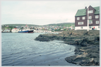 Building on rocks by water, with town on hillside beyond.