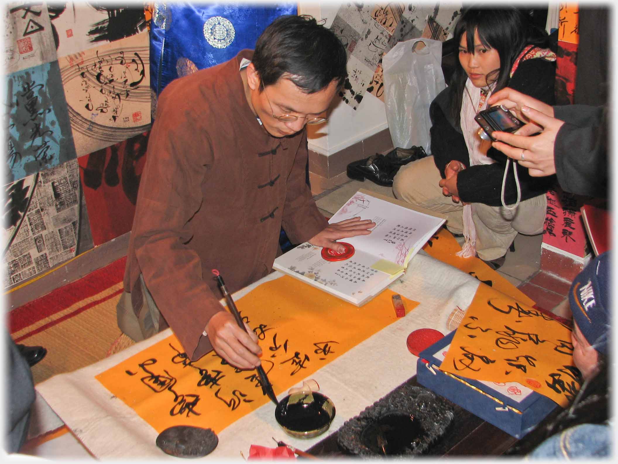 Man in brown tunic dipping a brush across a sheet with characters on it.