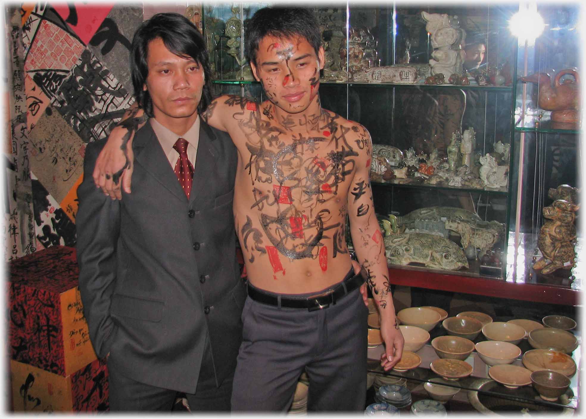 Man with torso covered in ink markings, arm on another man in suit's shoulders.