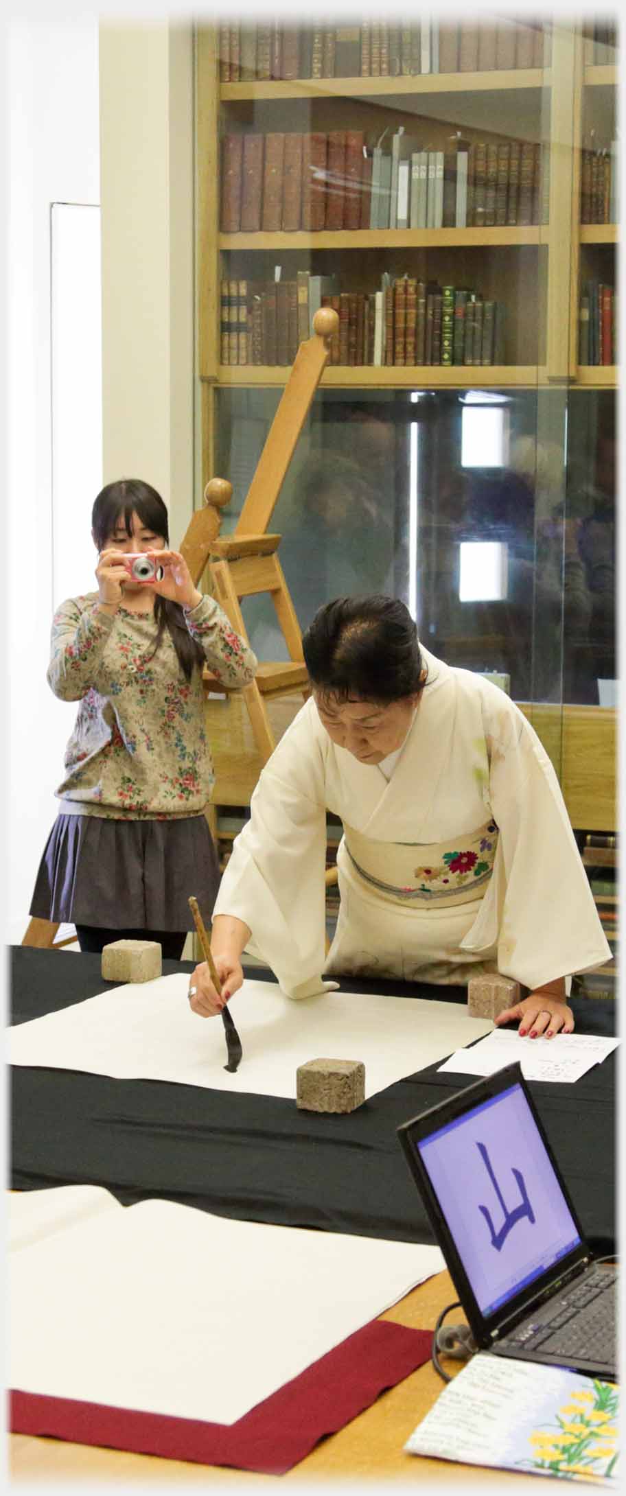 Kimono clad woman with paintbrush starting to write, woman  pbhotographing her.