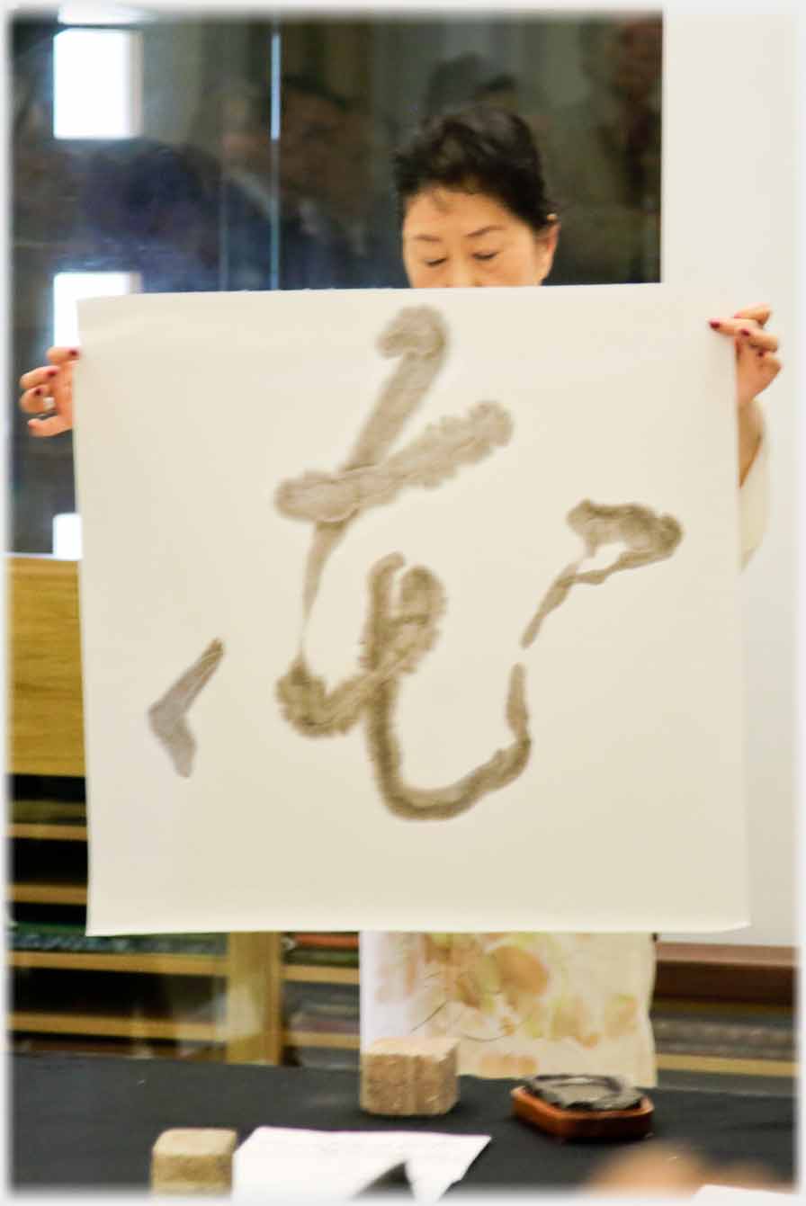 Woman in kimono holding up large sheet of paper with character on it.