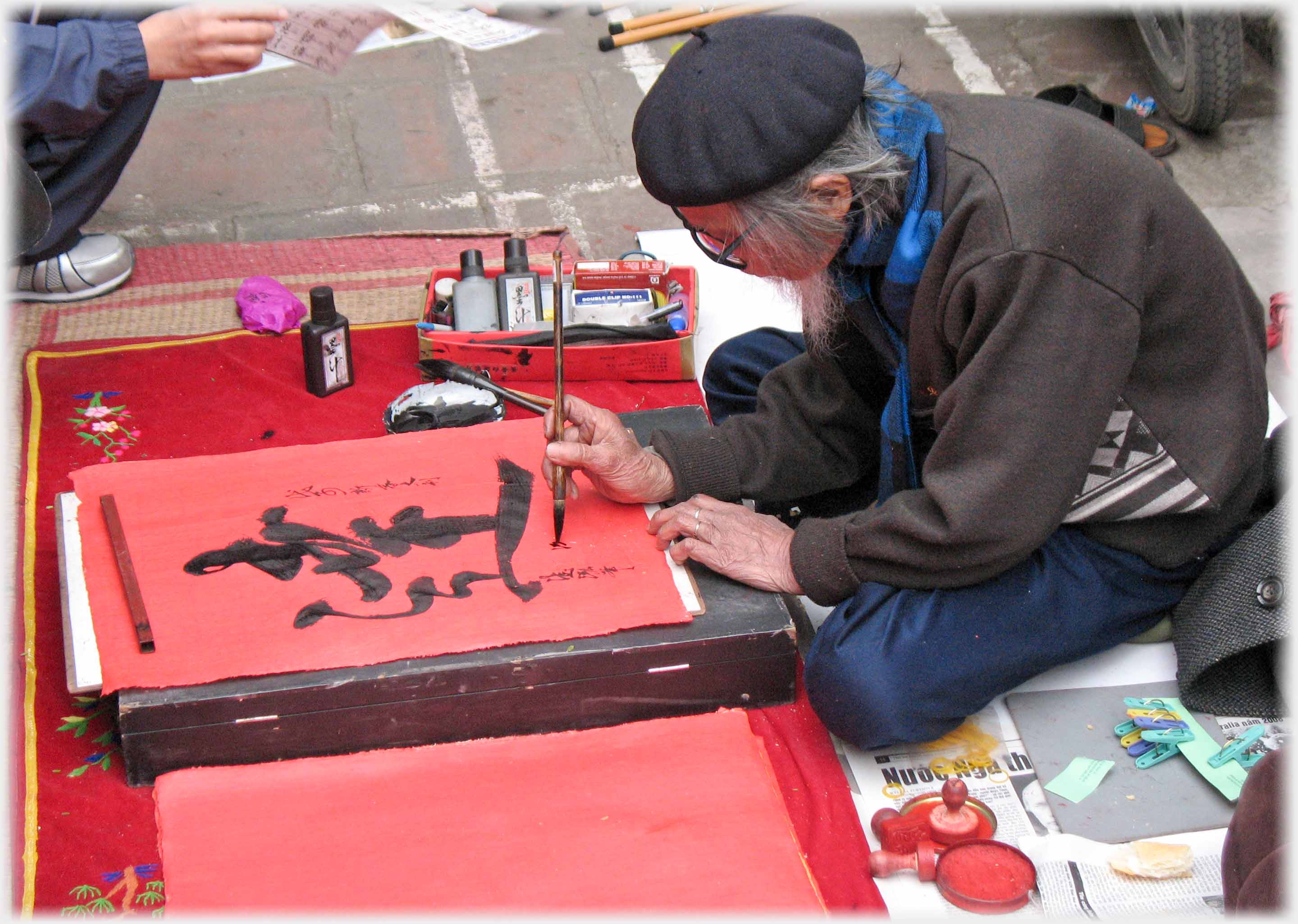 Man in beret cross legged on pavement writing Chinese characters with brush.