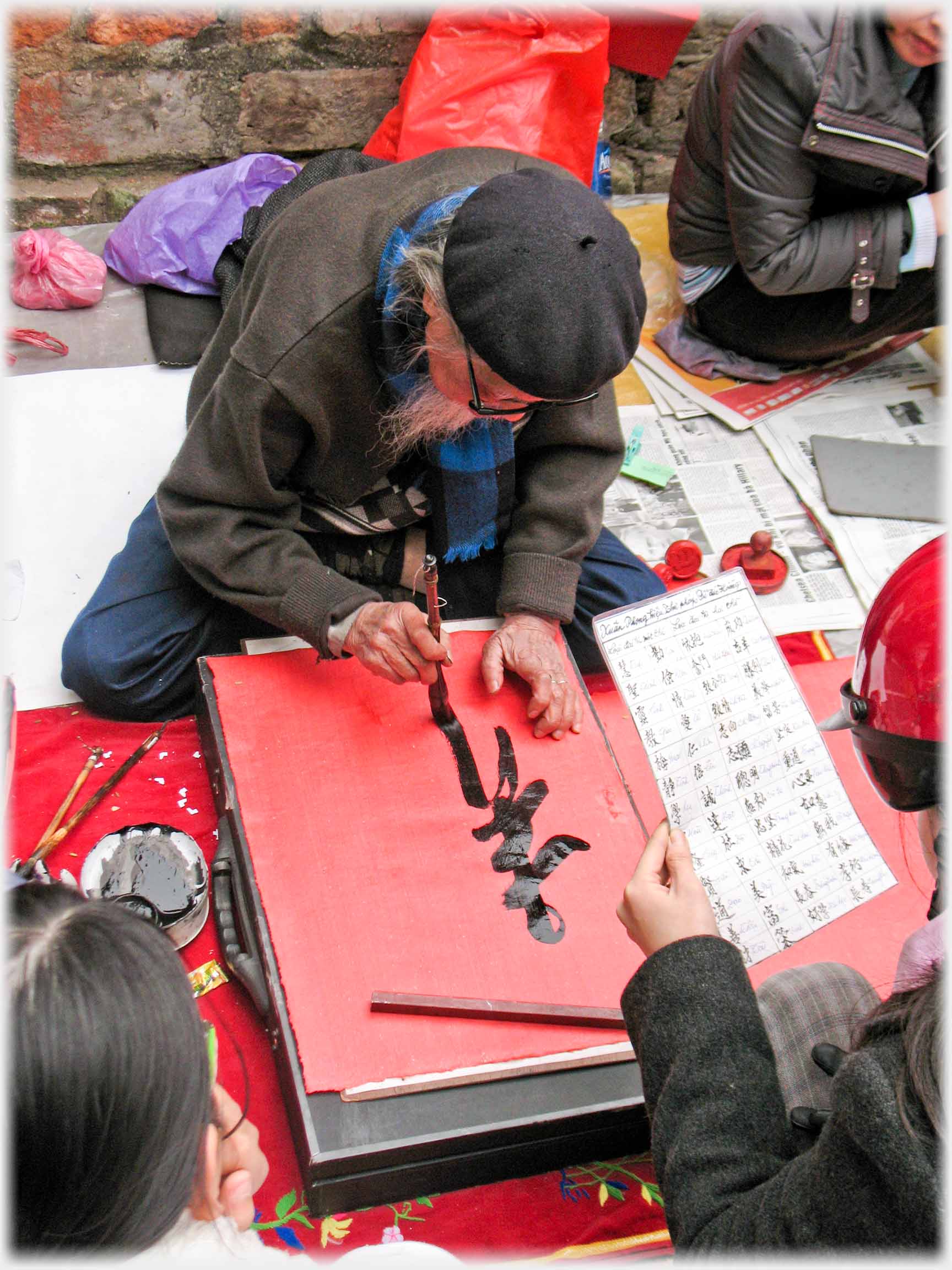 Man in beret writing with brush, hand holding list of Chinese characters.