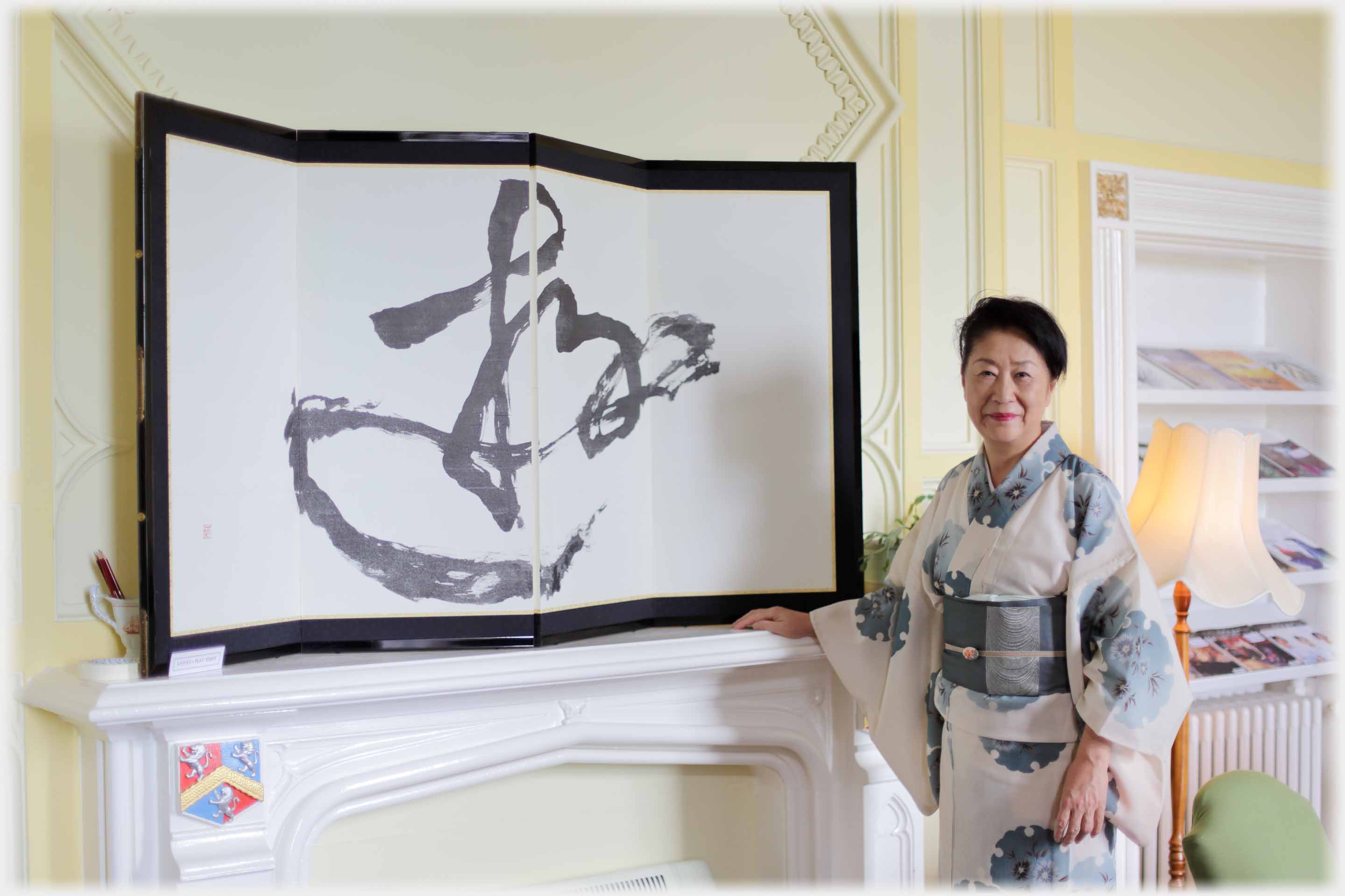 Woman in kimono standing with hand on mantlepiece on which there is a large Japanese calligraphic character.