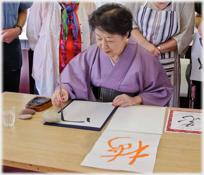 Six calligraphy class members standing around seated woman writing with brush.