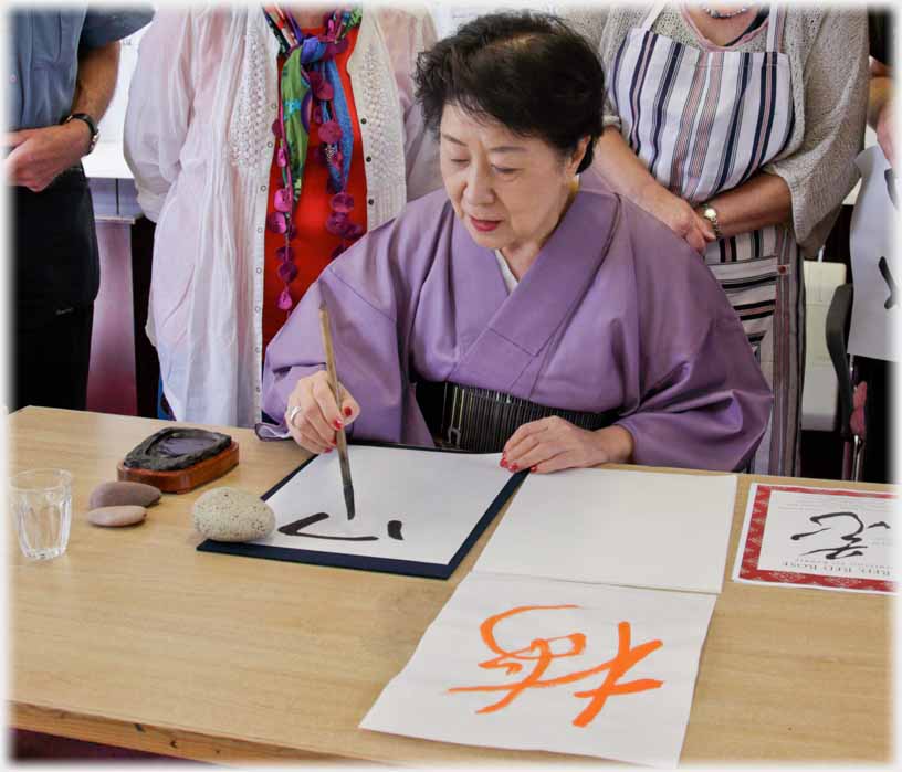 Six calligraphy class members standing around seated woman writing with brush.