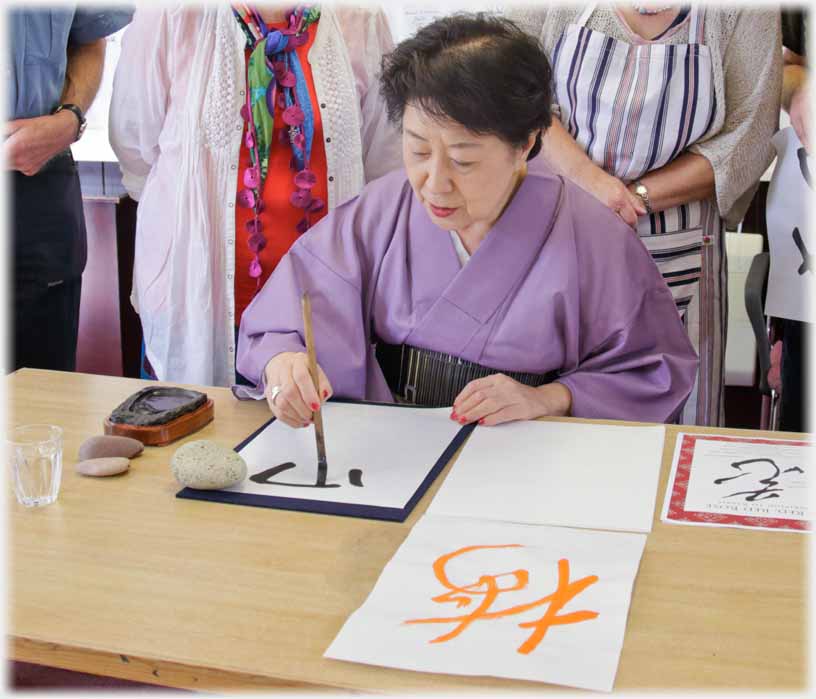 Six calligraphy class members standing around seated woman writing with brush.