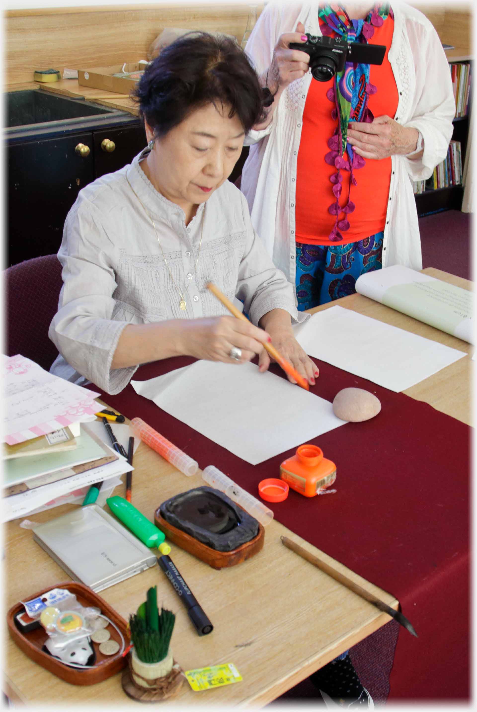 Woman sitting brush poised above blank paper, orange ink pot matching orange top of woman standing behind.