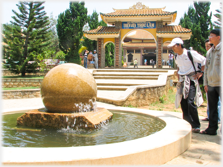 In a forecourt an aparent granite ball turns on water; people peering at it.