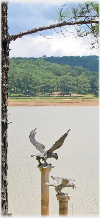 Two birds of prey, wings and beaks open on columns with lake in background.