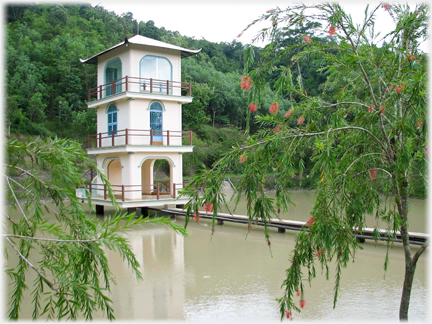 Three story square tower with windows and balconies in lake with pipe leading to it.