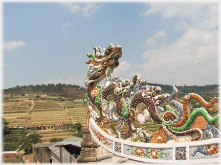 Brightly tiled dragon on the turned up corner of a roof looking back onto the roof with hills beyond.
