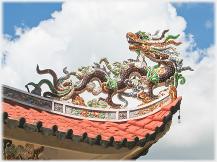 Brightly tiled dragon on the turned up corner of a roof looking back onto the roof with clouds above.