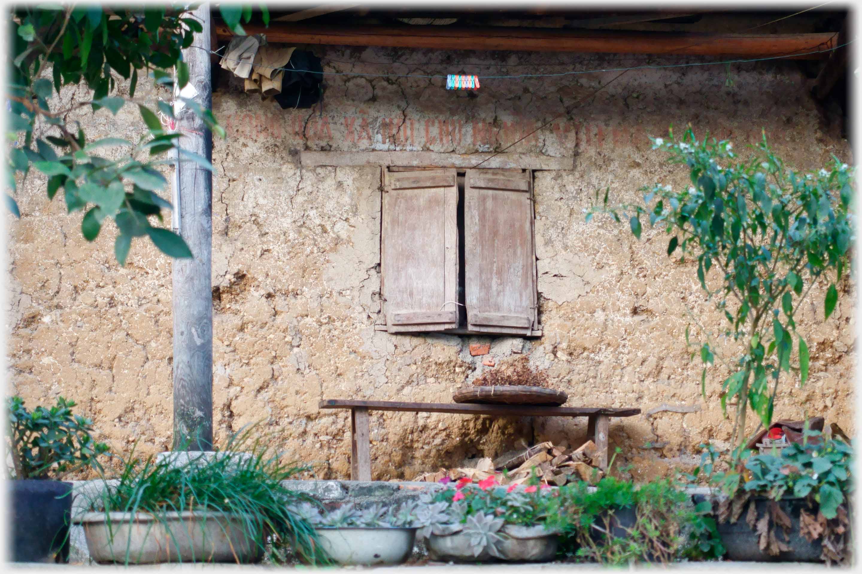 Closed shutters on small window in mudbrick wall with plants and seat.