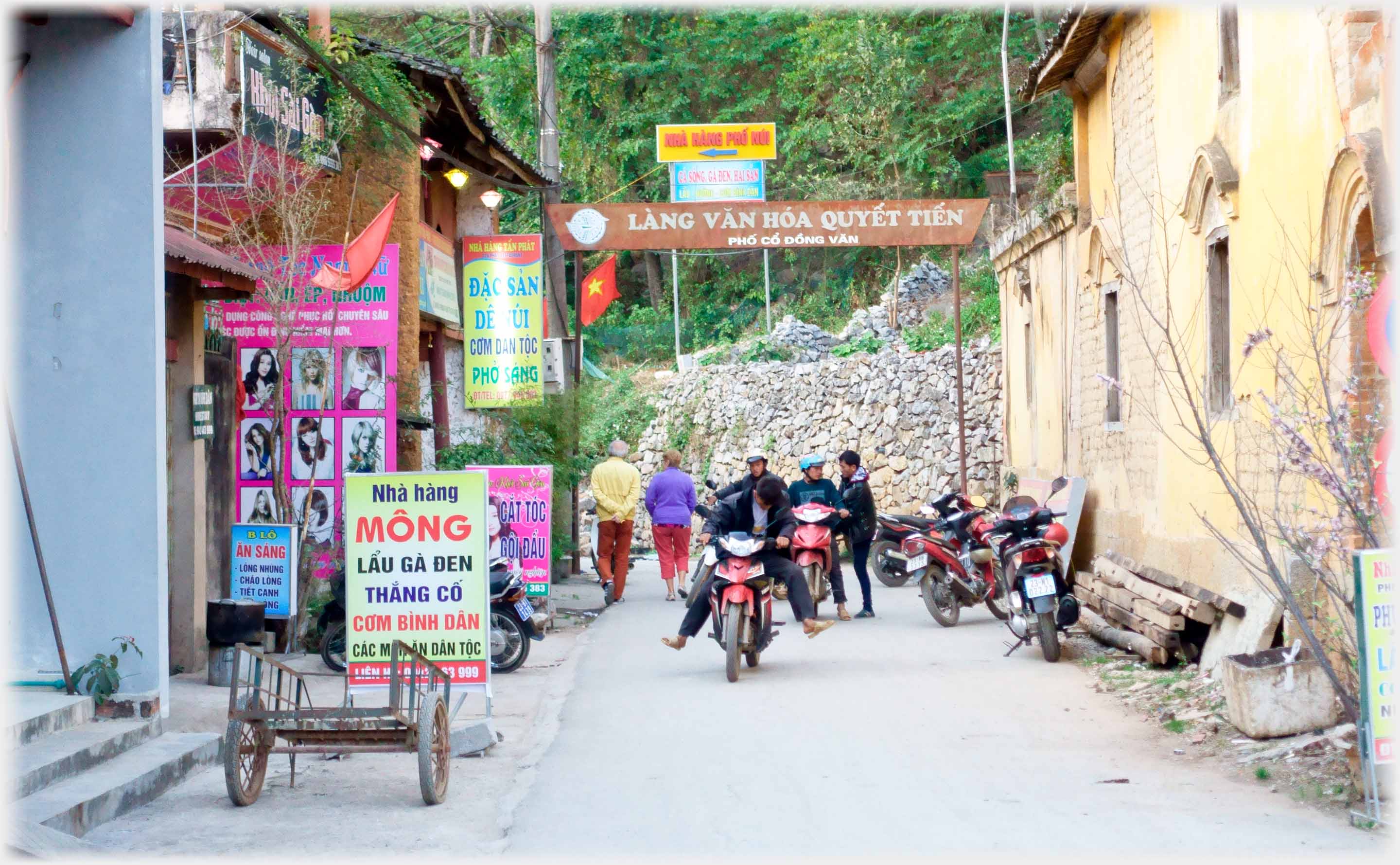 Oche building on one side, shops on other side iwth many signs, motorbikes moving and parked.