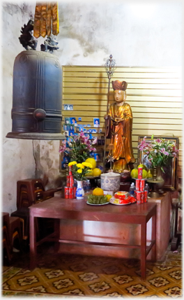 Small altar with statue and large bell beside it.