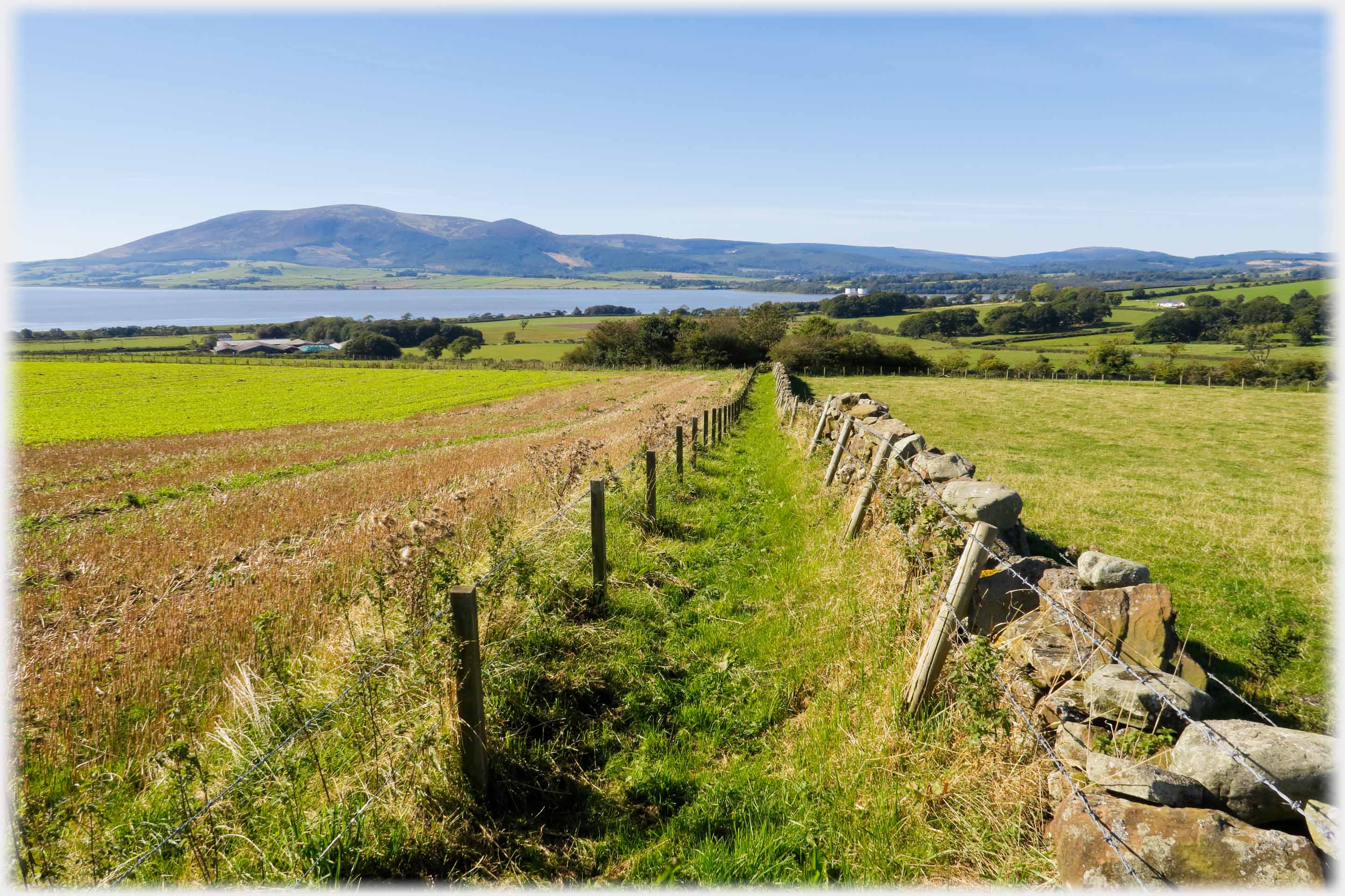 Path between dyke and fence running towards hill across water.
