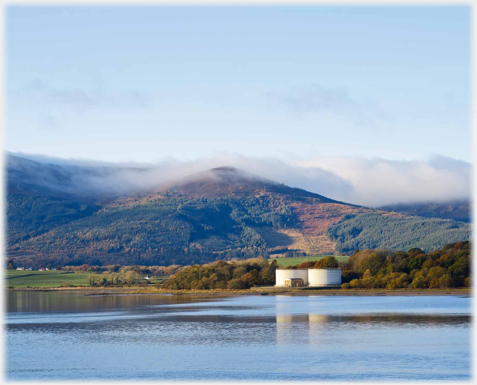 Small hill wreathed in cloud over two large white tanks.