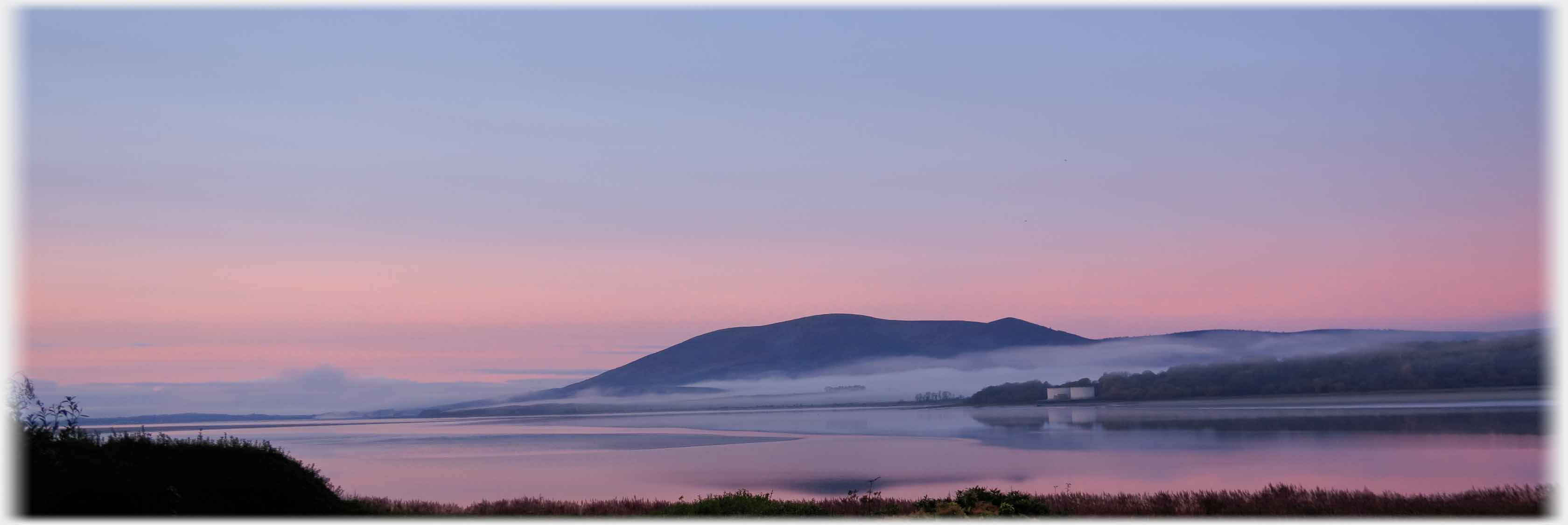 Pink lower sky and reflection, some mist hanging around hill.