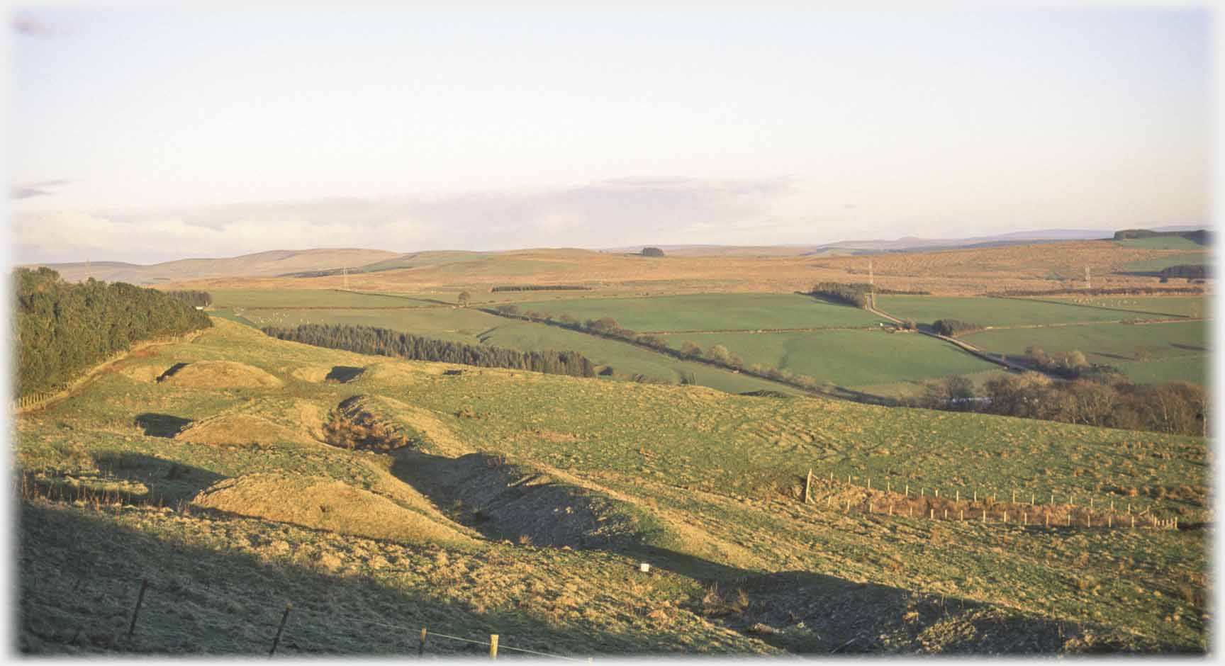 Three mounds with ditch and ridge beside them, open country.