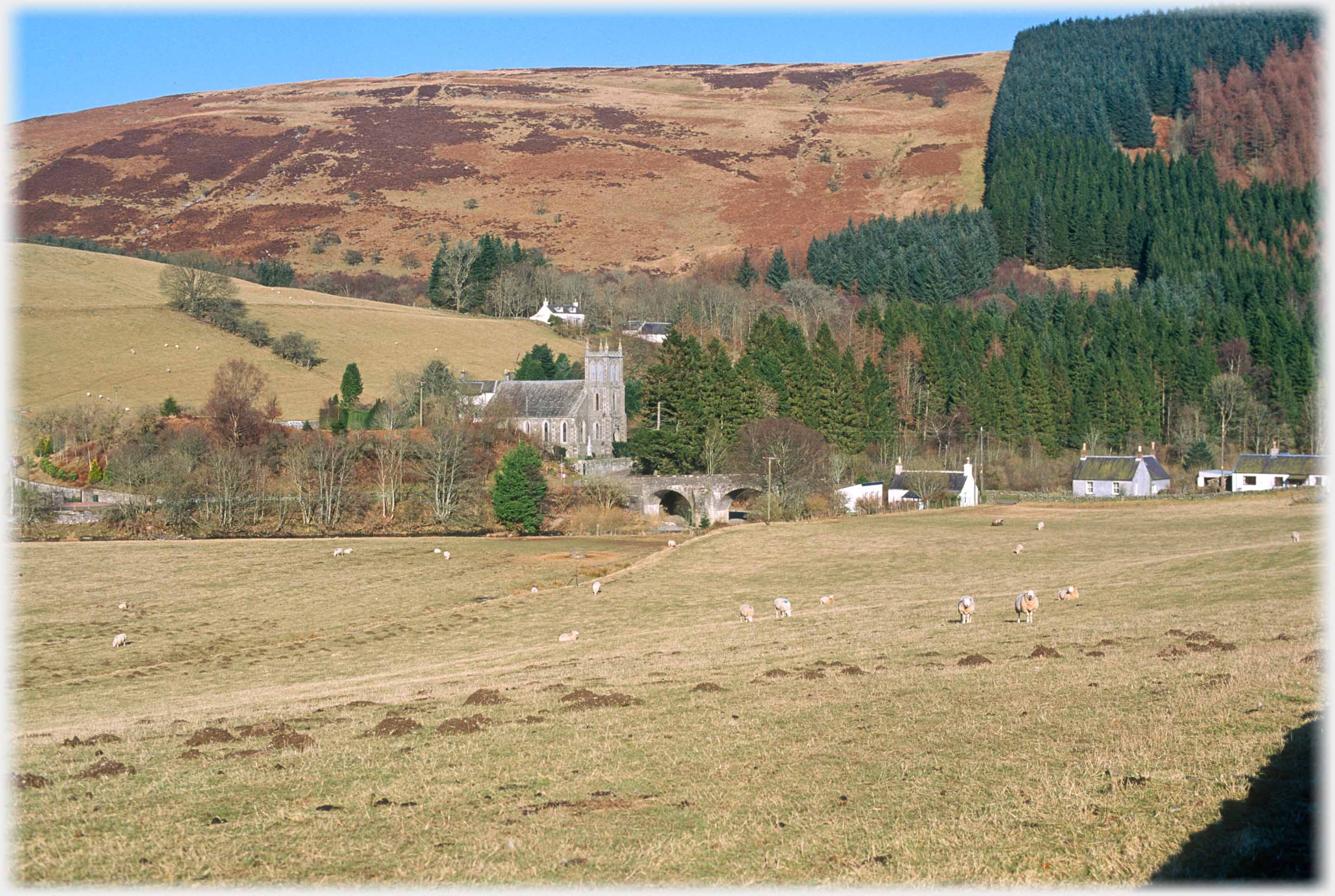 Church by bridge and trees, field with sheep in front, hill behind.
