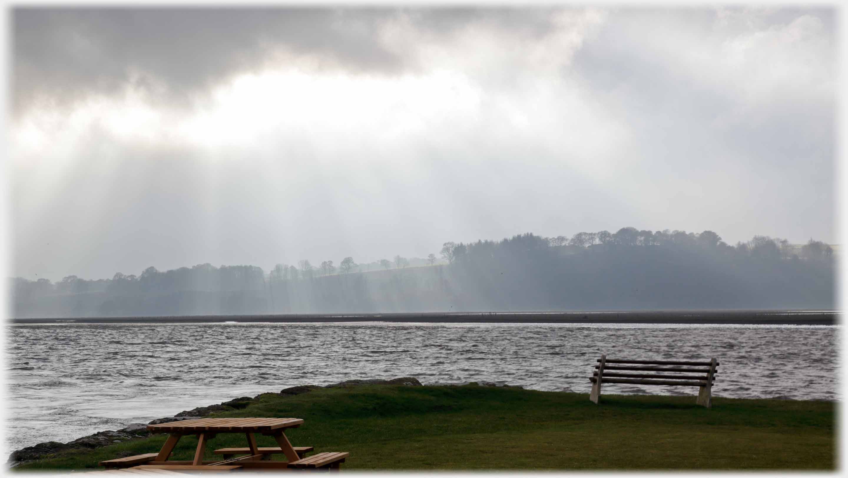 Seats overlooking water, towrds trees, with sun shafts from cloouds.
