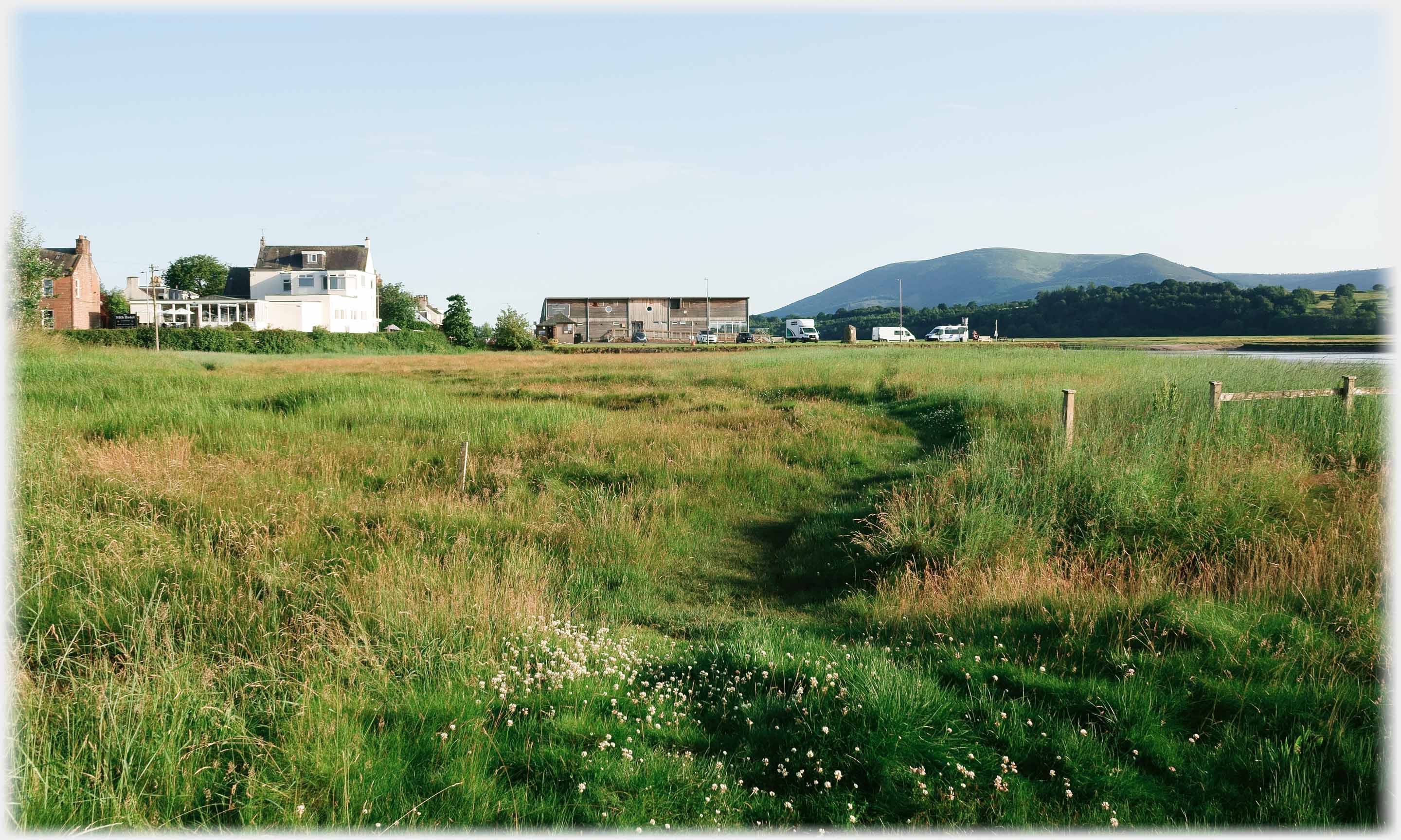 Curving path through grass towards two building and hill.