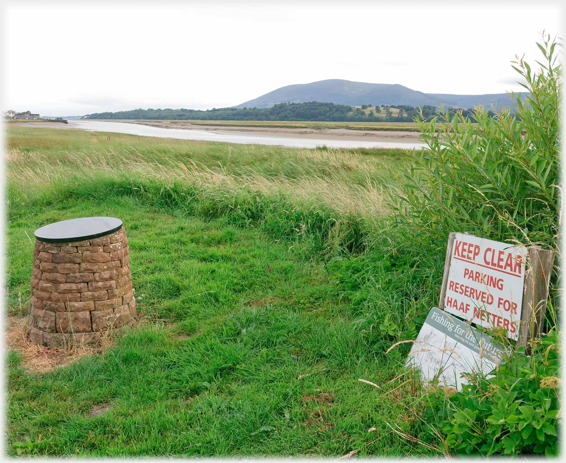Grassed area with stone cylinder and notice, river and hill beyond.