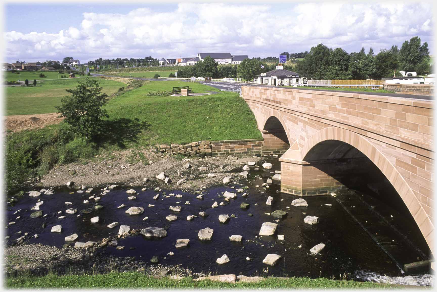 Red sandstone arched bridge with stones in shallow water below and houses beyond.
