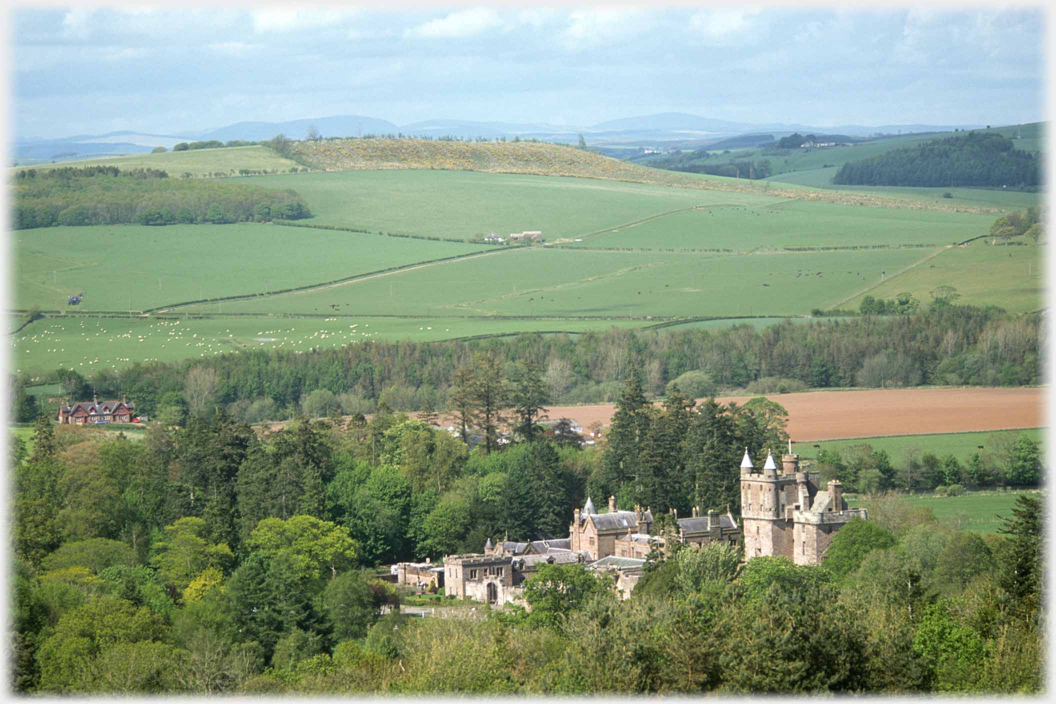 Tower and outbuildings among trees in valley, hills beyond.