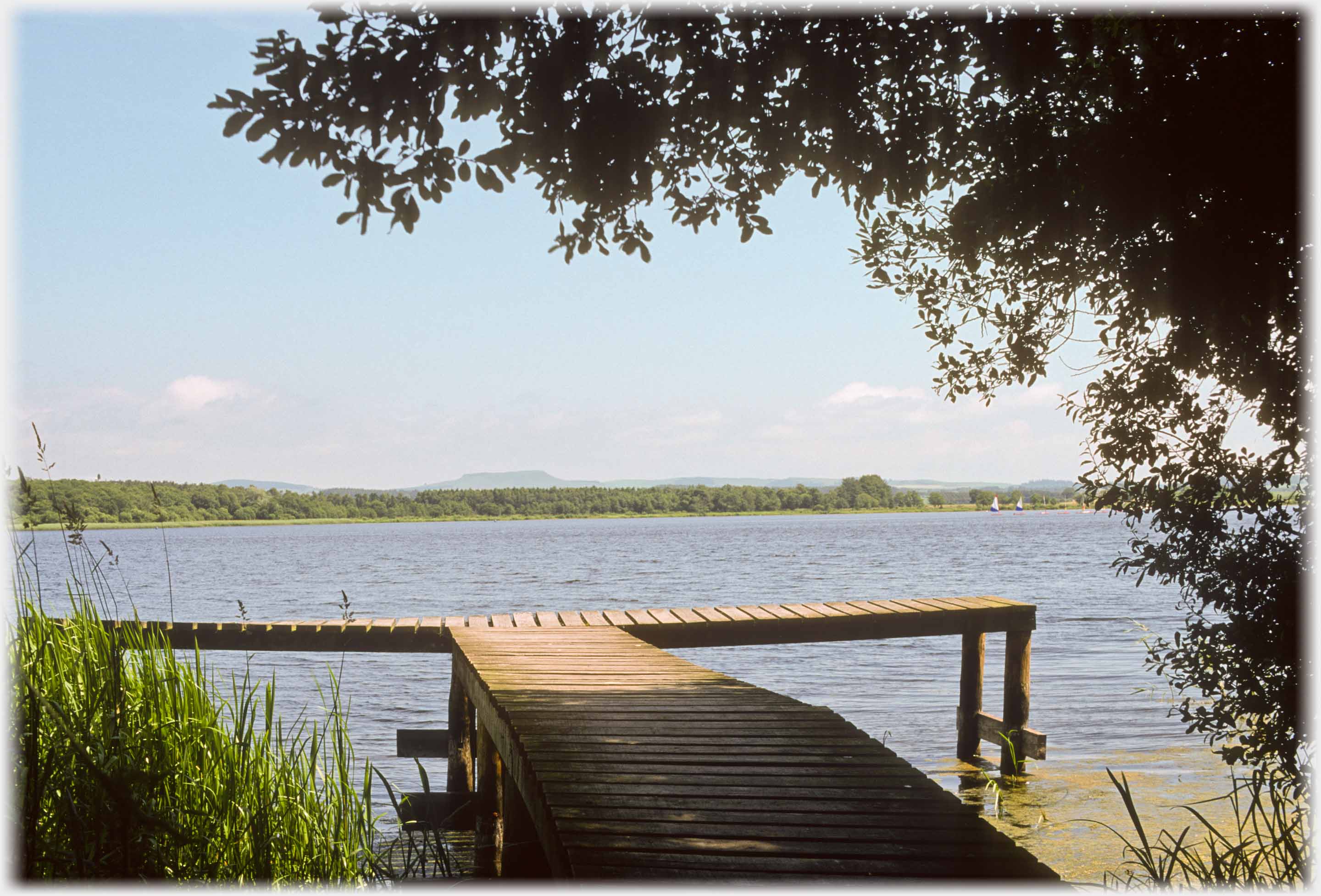 T-shaped jetty into large loch, flat hill in far distance.