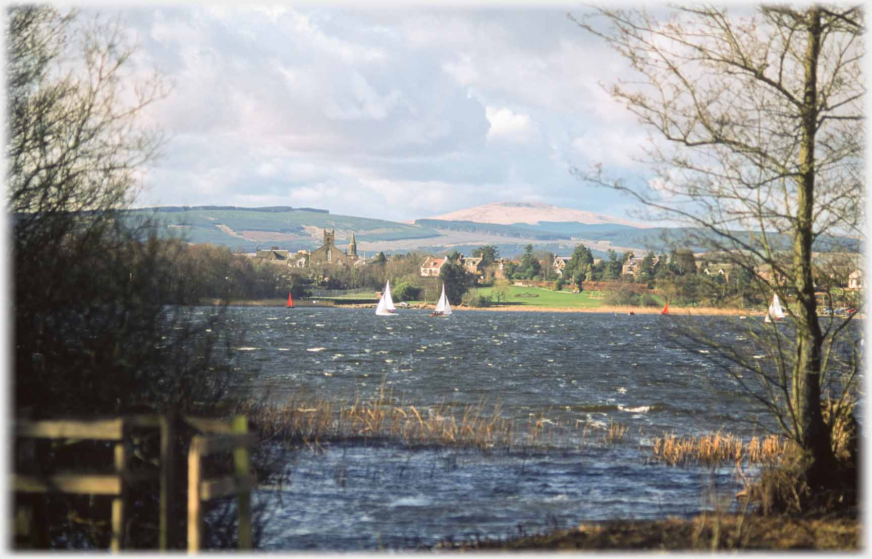 Slight waves on loch with sailing boats, village on far side.