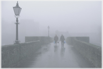 Two figures walking over bridge in mist.