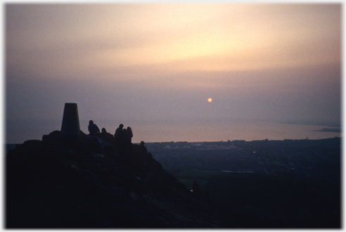 Silhouetted figures by a cairn with pin-point sun through clouds.