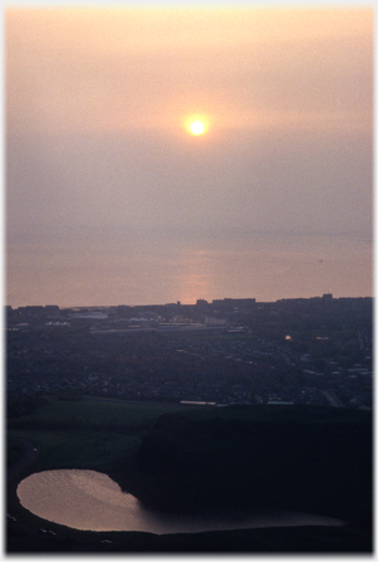 Sun over sea with buildings below and loch visible in the foreground.
