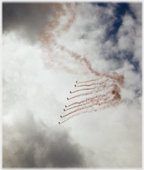 Team of sky divers with flares moving right-wards across sky.
