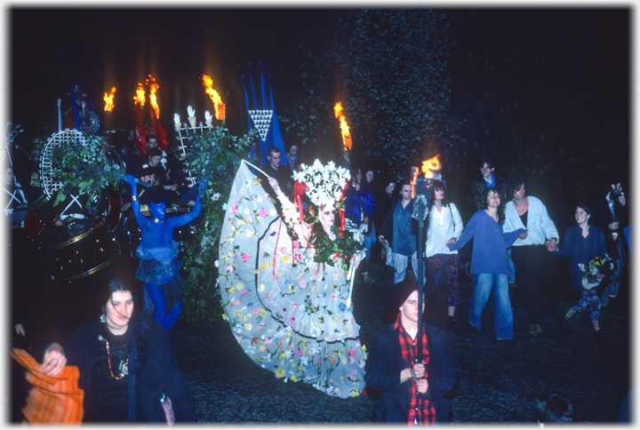 May Queen with headress and large flower studied dress in crowd.