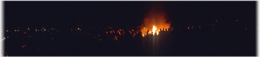 Wide panaramma of crowd, faces lit by small bonfire.