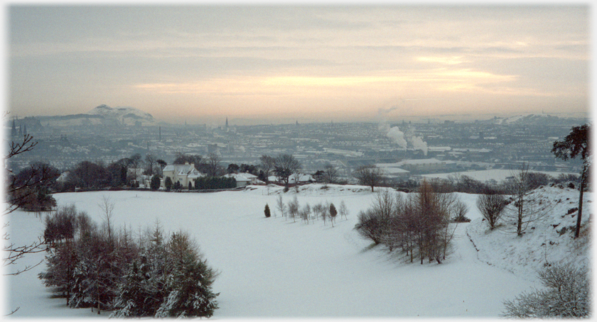 Snow covered golf coourse with city of white patches, smoking chimneys and clouds in warm sunrise.