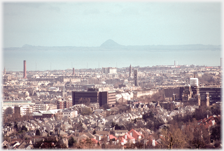 Rooftops of east of city with sea beyond and volcano shaped hill in centre of horizon.