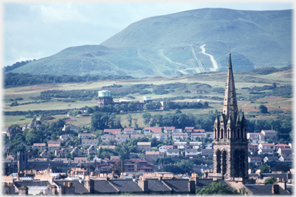 Church spire and rooftops iwth hills beyond and apparent roads on the hill.