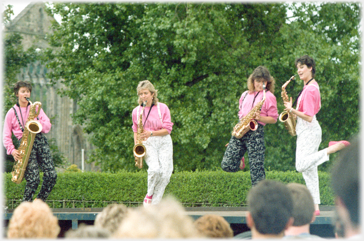 Four women harmoniously dressed jigging while playing saxaphones.