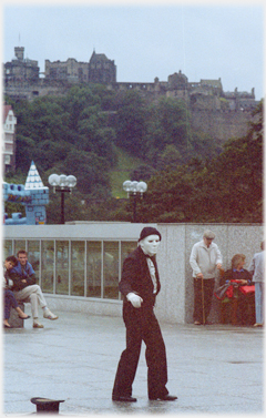Man in black clothes and white make up in low light with Castle beyond.