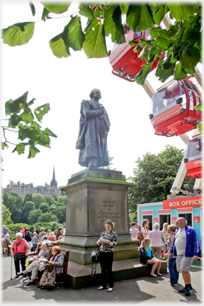 Statue on plinth with people all around and couple of ferris wheel gondolas in view.