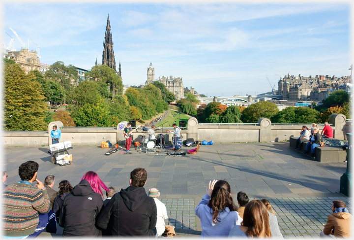 People sitting watching band performing with buildings beyond.