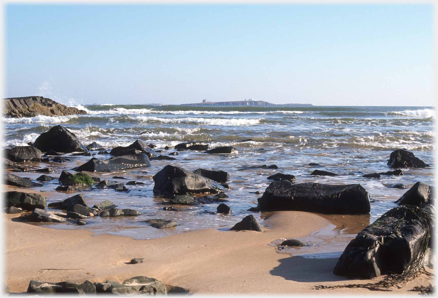 Rocks on sandy shore with waves and island beyond.