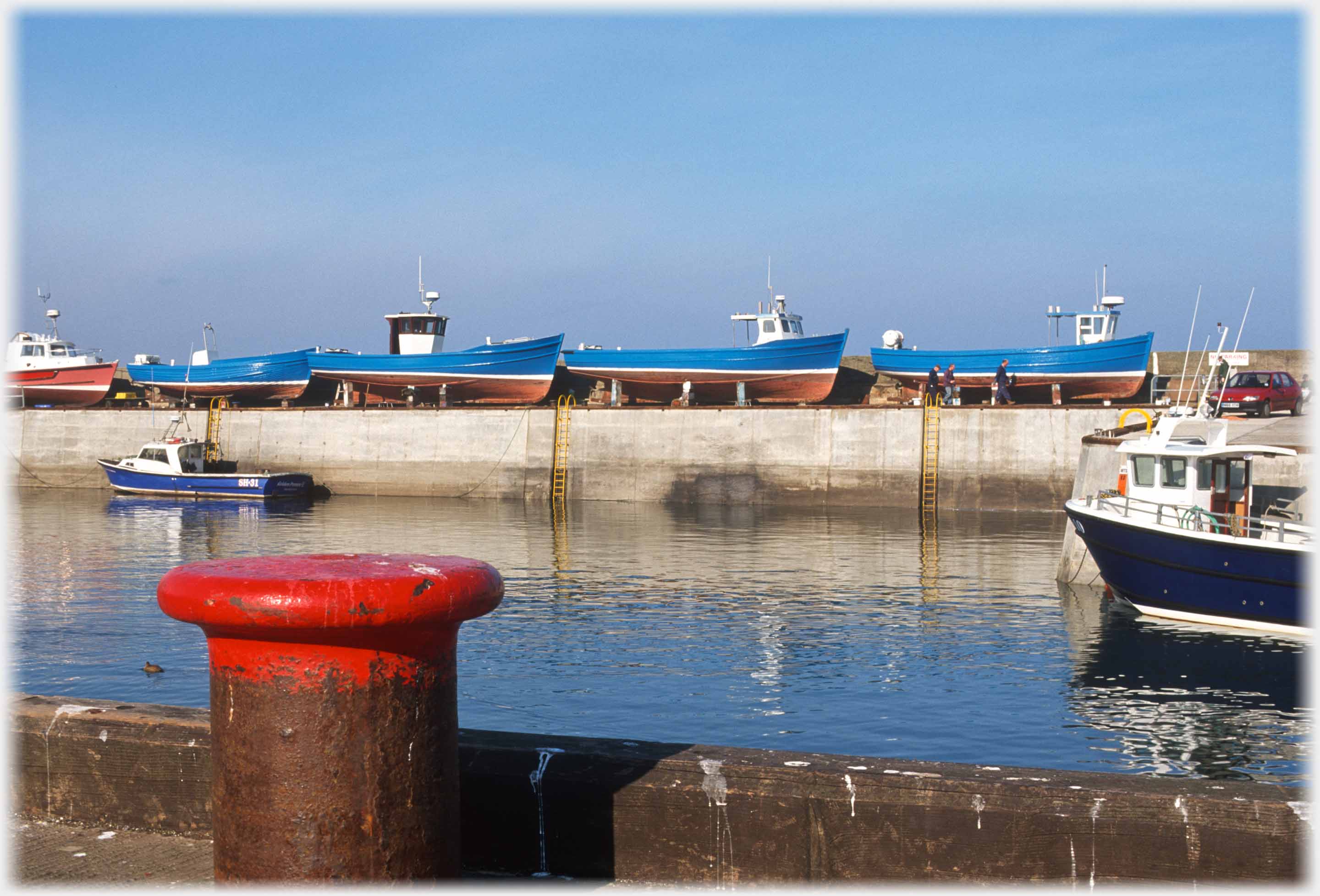 Red bollard by harbour with blue boats in background on quay.