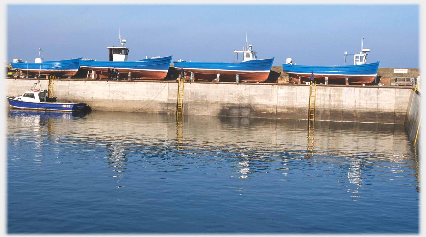 Three blue boats on quay with blue sky above and blue water below.