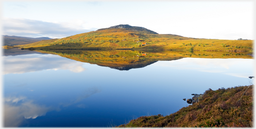 Small hill with house at its foot on the shore of a perfectly still blue loch.