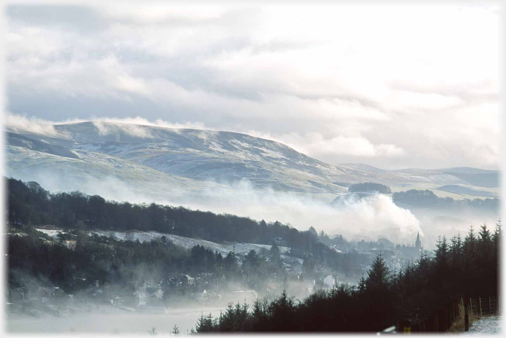 Billowing clouds in valley with hills and trees above.